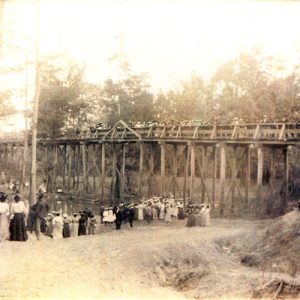 Group of people gathered at a river while spectators watch from a nearby wooden bridge