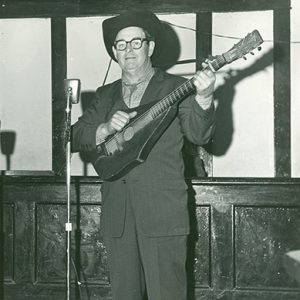 White man in suit and cowboy hat and glasses standing in front of a microphone stand and playing custom acoustic guitar