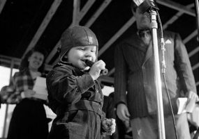 small child in leather helmet blows duck call at a microphone with adults standing in background
