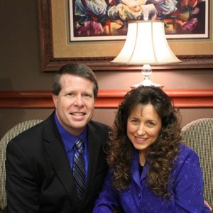 White man in suit and woman with long hair and purple shirt sitting in office chairs smiling