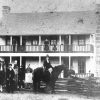 Various white people and two horses posing by large two-story building with chimneys on each end and wooden fence