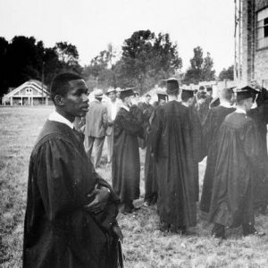 Black man in graduation gown outside brick stadium among graduates and people in suits dresses