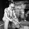 White man seated by stone hearth carving stick