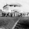 Group photo of black men women and children in formal attire outside multistory wood frame building