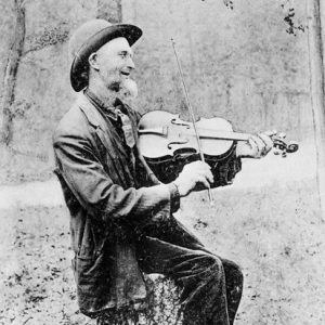 white man in suit and hat seated on tree stump playing fiddle, photo labeled "The Arkansas Fiddler"