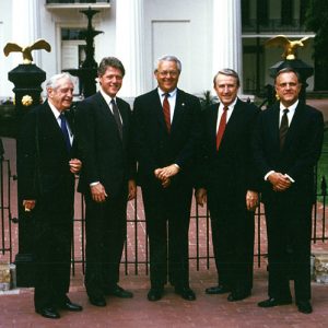 Group photo five white men in suits smile outside classical building brick walkway iron gate