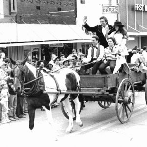 four white people in horse-drawn cart riding in parade down street lined with onlookers