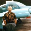 White man in pattern shirt and slacks posing by small airplane