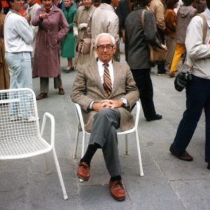 White man posing in suit seated among standing white crowd