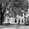 Two story brick home with small stoop in large tree shade by street parked car