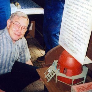 White man kneeling by small conical brick home model and text display on bench