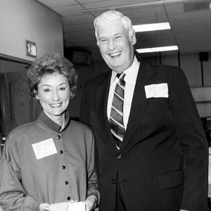 White woman button blouse and white man suit in name tags  pose smiling in office