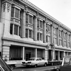 Three story stone building with ionic columns and bay windows and an entrance with an arch above it with cars parked on street in front