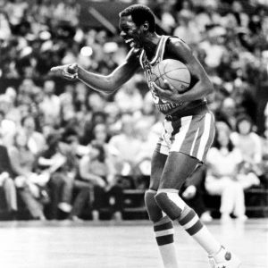 Black man in "Harlem Globetrotters" basketball uniform with ball on court smiling pointing with audience background