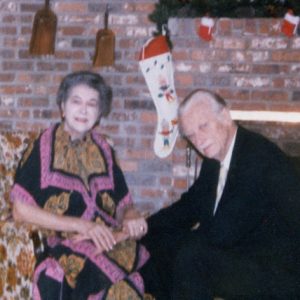 Elderly white woman in colorful dress and elderly man in suit seated holding hands by fireplace and Christmas stockings