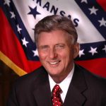 portrait white man smiling in suit and tie in front of Arkansas flag