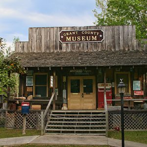 Single-story storefront building with covered porch and "Grant County Museum" sign under tree on grass