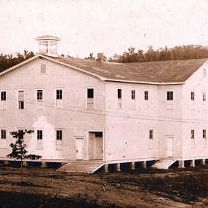 Multistory building with cupola on dirt lot with trees behind it