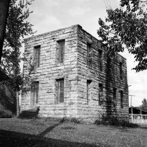 Austere two story stone building with window bars lawn trees nearby shed barn