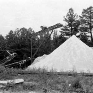 Large sediment pile by conveyor belt ramps tracks overgrown field metal building and tree line