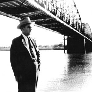 white man in suit and hat standing below bridge