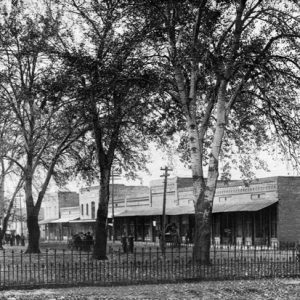 City square with trees pedestrians surrounded by iron fence and brick buildings horses and carts