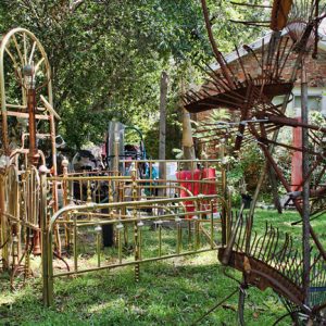 sculpture made from several brass headboards and footboards, and another made with rusted yard brooms, in front of brick house