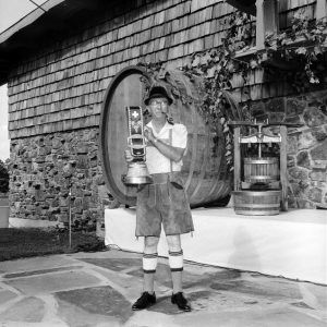 Older white man in lederhosen smoking pipe displaying large belt with bell outside wine cellar