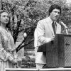 white woman gazes into crowd while white man speaks at lectern