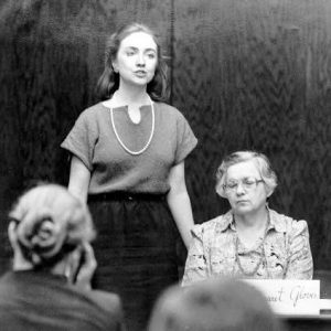 Young white woman in blouse skirt pearl necklace speaking to women committee and audience