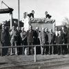 Group of white men in suits posing behind a rail with stone monument and building behind them