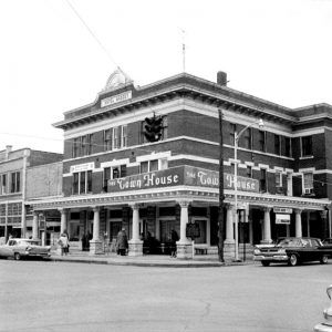 multistory brick building on corner downtown lot with "hotel massey" sign on the top and "the town house" sign on first floor with columned awning people are walking on sidewalk and cars are parked