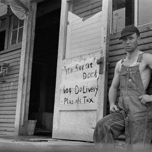 White man in cap and overalls standing outside his house with sign on open front door