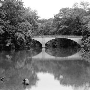 double arch support bridge over reflective forested river, half-submerged car wheel in shallow river foreground