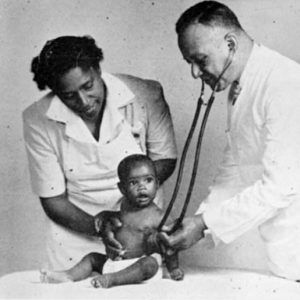 Black female nurse holding black infant on bed examined with stethoscope by black male doctor