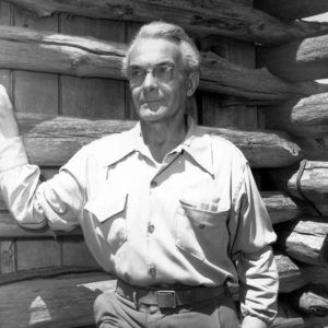 Portrait white man in outdoor wear, glasses, with hand on cabin wall exterior