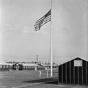 Flag raising with two soldiers, one saluting, in field near barracks.