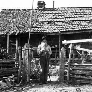 Old white man with beard standing outside cabin with covered porch and wooden fence