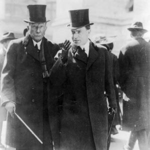 Two white men walking in suits overcoats top hats with canes on sidewalk with pedestrians