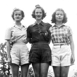 Three young white women in shirts and shorts standing with their arms around each other's backs