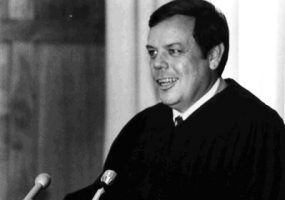 white man in black robe speaks into several microphones at lectern