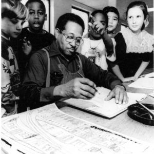 African-American man in overalls and glasses sitting at a table and drawing while surrounded by a mixed group of children