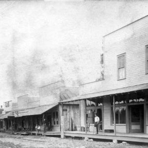 Dirt road through town with wood framed buildings and covered porches, man with one leg on crutches