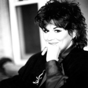 Portrait of a dark-haired white woman smiling with hand on chin with shaggy styled hair wearing jacket and lizard brooch