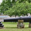 Side view of cannon balls and cannon on brick pedestals inside fence on grass with trees and buildings in the background