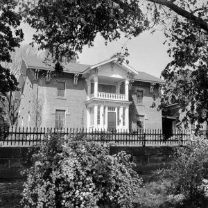 multistory brick home with stone lintels porch and balcony viewed over stone wall with iron fence and large bush in front