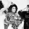 Black woman in white dress with bouquets being crowned by two white women also crowned