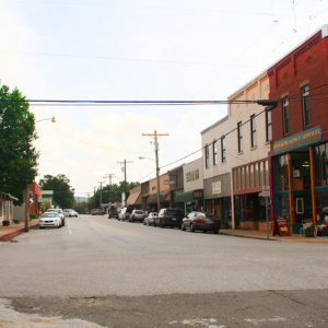 Parked cars outside of single and multistory brick storefronts on both sides of two-lane road