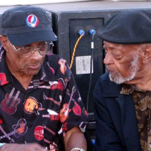 Two older black men in patterned collared shirts and caps sit by speakers conversing