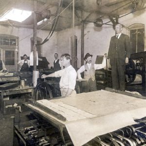 Old white man standing on a printing press while five white men are working in a print shop